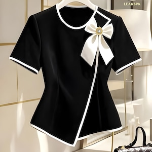 Può includere: Top nero a maniche corte con rifiniture bianche e un grande fiocco bianco con un accento di perla. Il top ha un design asimmetrico. I testi "LEAR" e "IMPORTED" sono visibili.