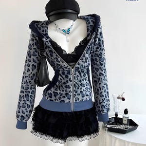 May include: A mannequin displays a gray and blue leopard print zip-up hoodie, black shorts, and a black cap. A black purse hangs on the mannequin's arm. A butterfly necklace and a black bra are visible. The text "LEAR" and "LEARSPK IMPORTED" are also visible.