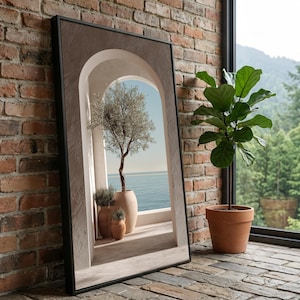 May include: A framed art print depicting an olive tree in a large beige pot on a balcony overlooking the sea. The print is in a black frame and leans against a brick wall. A potted plant sits nearby.