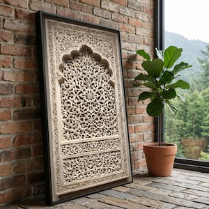 Op de afbeelding: Een ingelijst, crèmekleurig, ingewikkeld gesneden wandpaneel met een zwart frame. Het paneel heeft een gedetailleerd bloemen- en geometrisch ontwerp. Het leunt tegen een rode bakstenen muur, met een plant in een pot en een raam op de achtergrond.