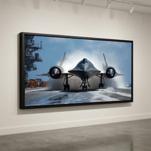 May include: A framed print of a black SR-71 Blackbird aircraft on a runway, with vapor rising from the plane. The image shows the aircraft's distinctive shape and the surrounding environment, including personnel in orange uniforms.