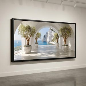 May include: A framed print depicts a scenic coastal view. White archways frame a pathway lined with potted trees, leading to a view of the sea and a large white cliff. A sailboat is visible in the distance. The frame is black.