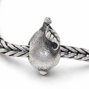May include: A silver charm bead with a pear-shaped design, featuring a textured surface and a small leaf detail. The bead is strung on a silver chain, creating a jewelry piece. The charm is approximately 0.5 inches in length.
