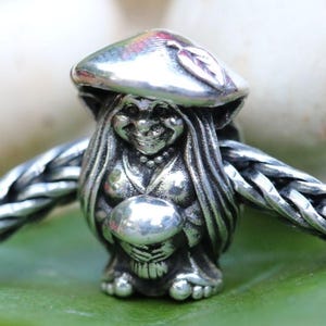 May include: A silver charm depicting a whimsical figure with long hair and a mushroom-shaped hat. The charm is holding a sphere and is attached to a braided silver chain. The charm is resting on a green leaf.