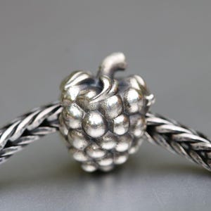 Op de afbeelding: Een zilveren bedel in de vorm van een framboos, met een getextureerd oppervlak en steel. De bedel is geregen aan een zilveren gevlochten ketting. Het is een sieradencomponent.