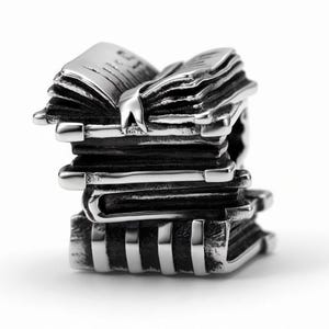 May include: A silver-toned metal charm shaped like a stack of four books. The top book is open, revealing pages and a bookmark. The metal has a polished finish on the edges and a darkened, oxidized texture in the recessed areas of the book covers.