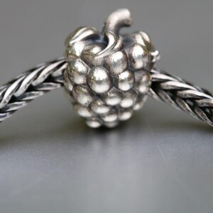 Op de afbeelding: Een zilveren bedel in de vorm van een framboos, geregen aan een gevlochten zilveren ketting. De bedel heeft een gedetailleerd, getextureerd oppervlak met kleine, ronde bobbels. De ketting is ook zilver en heeft een geweven uiterlijk. De achtergrond is effen grijs.