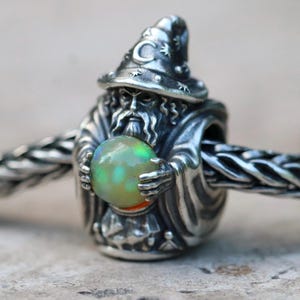 May include: A silver bead charm shaped like a wizard wearing a pointed hat with a crescent moon and star motif. The wizard figure holds a round, iridescent opal sphere between both hands. The charm is threaded onto a silver chain link bracelet.