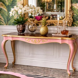 May include: A pink and gold console table with a dark wood top. The table features ornate gold accents and curved legs. Decorative items on the table include vases, a golden pineapple, and glassware. The background includes a mirror and floral wallpaper.