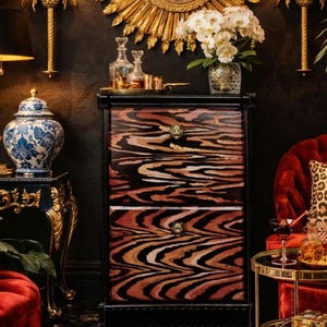 May include: A two-drawer wooden chest featuring a black frame and drawer fronts with a wavy, multi-toned brown and black tiger-stripe pattern. Each drawer is equipped with a small, metallic gold-toned tiger head handle centered on the front.