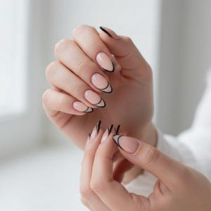 May include: Close-up of a hand with almond-shaped nails. The nails feature a nude base with a thin black outline, a modern French manicure. The nails are long and well-manicured, showcasing a minimalist nail art style.