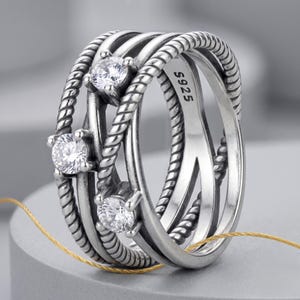 May include: A silver ring featuring three clear, round gemstones. The ring has a multi-band design with a twisted rope texture on some bands. The inscription "S925" is visible on the ring's inner band. A piece of jewellery.