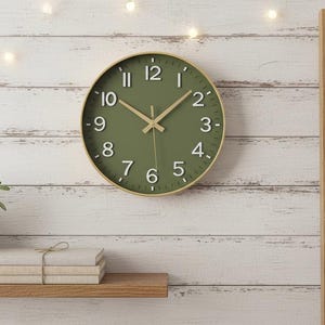 May include: A round olive green wall clock with white numerals and gold-colored hands. The clock is mounted on a white wooden wall. A small potted plant, books, and a blanket on a wooden ladder are also visible.