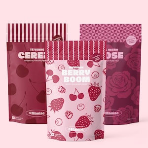 Può includere: Tre confezioni di tè con disegni di bacche e fiori. La confezione centrale recita "Berry Boom Infusion" con illustrazioni di fragole, mirtilli e ciliegie. Le altre confezioni presentano disegni di ciliegie e rose. Il marchio "Maniac" è visibile.