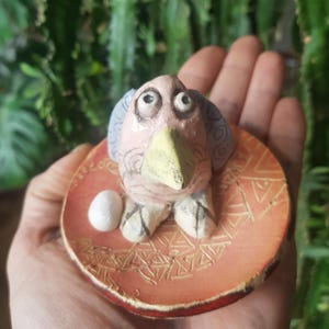 Può includere: Figurina di uccello in ceramica dipinta a mano, con becco giallo, posata su una base rotonda arancione. La base presenta un motivo geometrico e un piccolo uovo bianco. L'uccello ha un corpo rosa e dettagli blu.