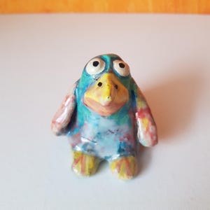 Könnte beinhalten: Handbemalte Keramik-Pinguin-Figur. Der Pinguin hat einen blauen Kopf, einen gelben Schnabel und einen weißen Bauch mit farbenfrohen Akzenten in Rot, Rosa und Gelb. Die Füße des Pinguins sind gelb mit orangefarbenen Streifen.