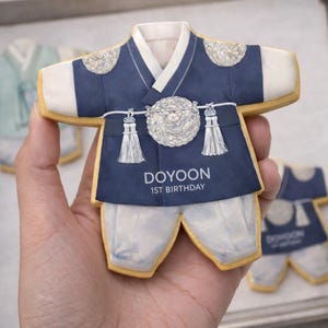 May include: A cookie shaped like a traditional Korean hanbok, in navy blue, white, and beige. The cookie has a detailed design, including a decorative silver emblem and the text "DOYOON 1ST BIRTHDAY".
