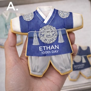 May include: A cookie shaped like a traditional Korean hanbok in blue and white. The design features a circular silver emblem, tassels, and the printed text ETHAN 100th DAY. The garment has white sleeves, a blue bodice, and white trousers.