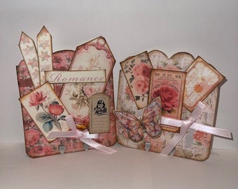 May include: Two pink and white papercraft boxes decorated with floral patterns, butterflies, and ribbons. The boxes have the word "Romance" printed on one of them.