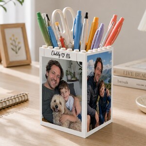 Peut inclure: Un pot à crayons blanc en forme de bloc, orné de photos d'un homme et d'un enfant. Il contient des stylos, des ciseaux et des articles de papeterie. Le texte "Daddy ♥ Me" est imprimé.