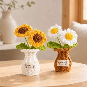Puede incluir: Dos arreglos florales de ganchillo en jarrones pequeños. Un jarrón blanco contiene dos girasoles amarillos con centros marrones. Un jarrón marrón contiene dos margaritas blancas con centros amarillos. Cada jarrón tiene una etiqueta Handmade with love.