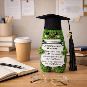 May include: A green crocheted graduation pickle wearing a black mortarboard and holding a sign that reads "Congratulations, Graduate!" The tassel is black with a gold 2026 charm. The sign has a jar design.