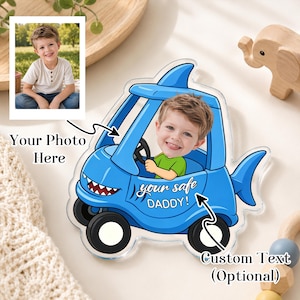 Peut inclure: Une voiture requin personnalisée en acrylique avec la photo d'un enfant au volant. La voiture bleue a des roues noires et le texte "your safe DADDY!". L'image comprend un cadre photo avec le texte "Your Photo Here" et "Custom Text (Optional)".