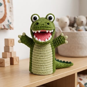Puede incluir: Una marioneta de mano de ganchillo con forma de cocodrilo verde con vientre verde claro, una boca muy abierta con dientes blancos y un interior rosa, ojos grandes en blanco y negro y una cola texturizada que sale de la parte posterior.
