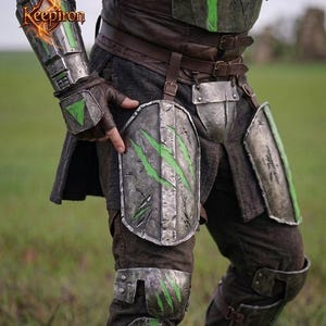 Puede incluir: Primer plano de una armadura de guerrero, con placas de metal desgastado y detalles en verde. La armadura incluye protectores de brazos, protecci&oacute;n para las piernas y una pieza para el pecho, todo asegurado con correas de cuero. El logo "Keepiron" es visible.