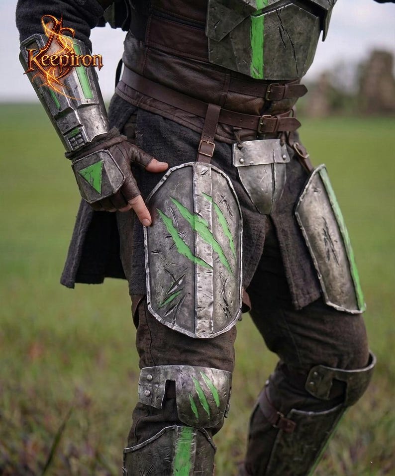 Puede incluir: Primer plano de una armadura de guerrero, con placas de metal desgastado y detalles en verde. La armadura incluye protectores de brazos, protecci&oacute;n para las piernas y una pieza para el pecho, todo asegurado con correas de cuero. El logo "Keepiron" es visible.