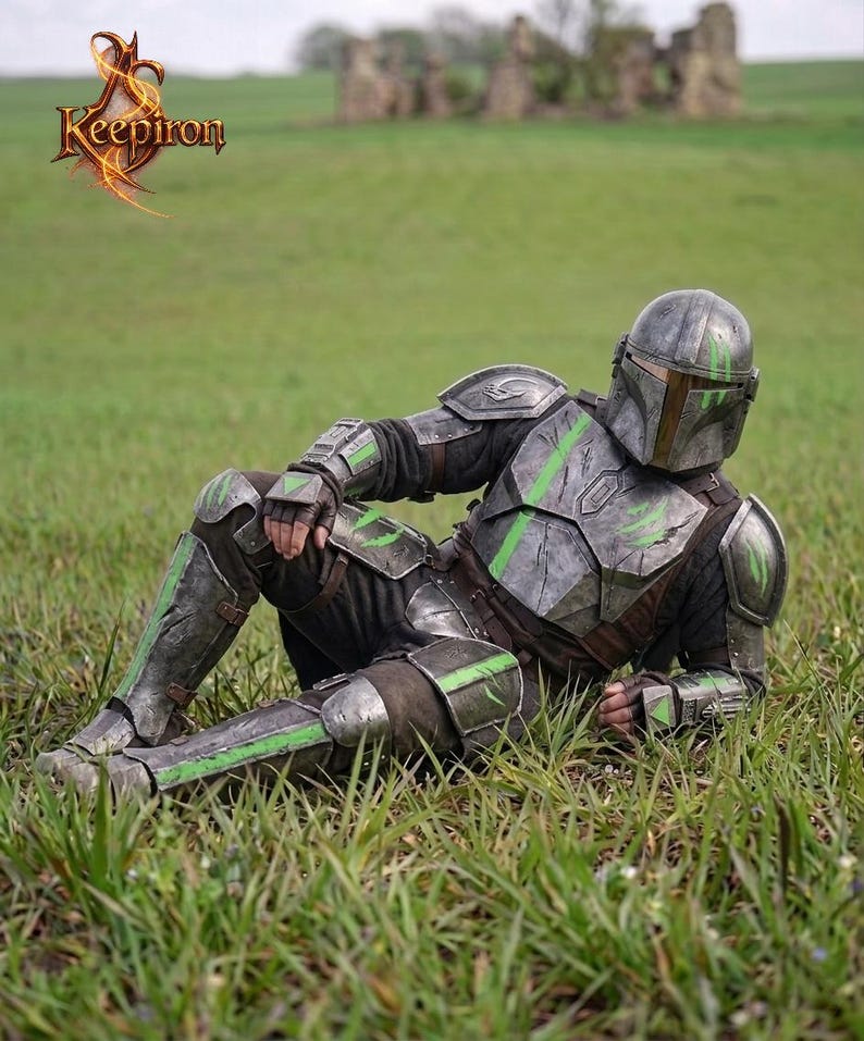 Puede incluir: Una persona con una armadura detallada, gris met&aacute;lico y verde, casco y guantes, descansando en un campo de hierba. La armadura tiene detalles en verde y est&aacute; dise&ntilde;ada con detalles intrincados. El logotipo "Keepiron" es visible en la esquina superior izquierda.