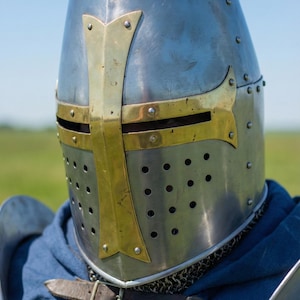 May include: A silver-colored knight's helmet with a gold cross design on the front. The helmet has small circular ventilation holes and is worn with a blue garment and chainmail.