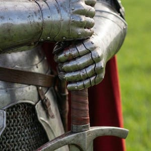 Pode incluir: Close-up de uma espada medieval segurada por uma pessoa em armadura completa. A espada tem uma guarda em forma de cruz e uma empunhadura envolta em couro. A armadura &eacute; prateada e mostra sinais de desgaste. Uma capa vermelha &eacute; vis&iacute;vel.