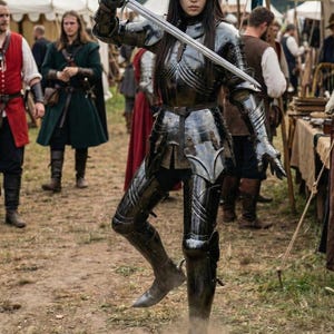 May include: A person in full black armor, holding a sword aloft. The armor is highly polished, reflecting the light. The scene appears to be at a medieval fair or reenactment, with other costumed people in the background.