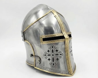 May include: A silver-colored medieval helmet with gold-colored trim and a cross design. The helmet has a rounded top and a face guard with a horizontal eye slit. The gold trim features intricate patterns.