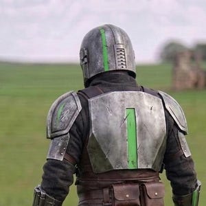 Puede incluir: Una persona con una armadura plateada y verde, vista desde atr&aacute;s, en un campo. El casco y la placa del pecho tienen detalles verdes. El fondo muestra un campo verde y un cielo nublado.