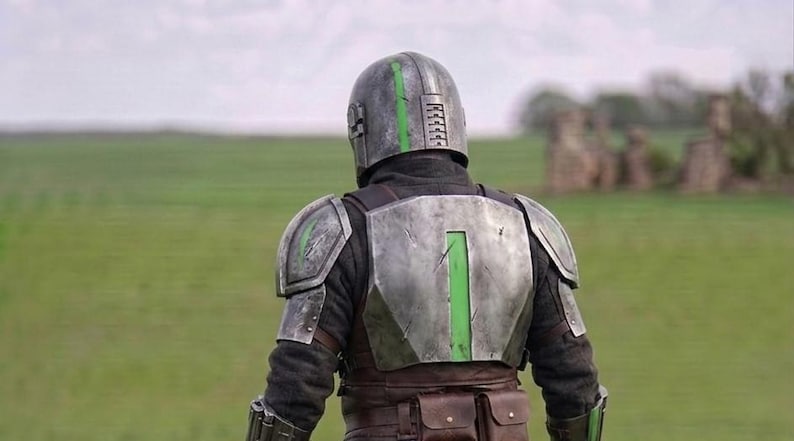 Puede incluir: Una persona con una armadura plateada y verde, vista desde atr&aacute;s, en un campo. El casco y la placa del pecho tienen detalles verdes. El fondo muestra un campo verde y un cielo nublado.