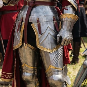 May include: Close-up of medieval armor, featuring polished metal plates with gold trim. A burgundy leather belt and accents add color. The armor includes a gauntlet, leg protection, and a chest plate, suggesting a knight's attire.