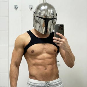 May include: A polished, silver Mandalorian helmet with a black visor. The person is wearing a black harness-style top and grey sweatpants. The person is holding a phone. The helmet is the main focus of the image.