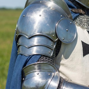 May include: Close-up of polished steel armor, including shoulder and elbow protection, with rivets. A blue cape is visible, along with chainmail and a white garment with a black cross design. The armor reflects the sunlight.