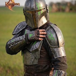 Puede incluir: Casco y armadura mandalorianos detallados en tonos grises, marrones y verdes. La armadura presenta detalles desgastados por la batalla y acentos verdes. El logotipo de Keepiron es visible en la esquina superior izquierda. El tema general es cosplay y ciencia ficci&oacute;n.