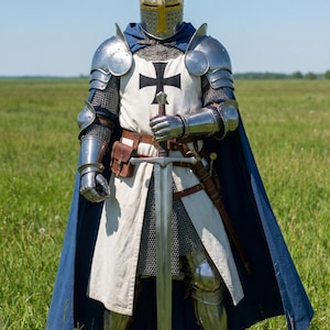 May include: A full suit of medieval armor, including a helmet, shoulder guards, and leg protection. The armor is silver-colored, with a white tunic featuring a black cross. A blue cape and a long sword complete the ensemble. The person is standing in a grassy field.