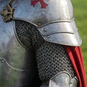 Pode incluir: Close-up de uma armadura de cavaleiro medieval. A armadura de metal prateado apresenta um emblema de cruz vermelha na ombreira. Cota de malha e uma capa vermelha tamb&eacute;m s&atilde;o vis&iacute;veis, sugerindo um tema hist&oacute;rico ou de fantasia.