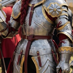 May include: A detailed view of medieval armor, showcasing polished metal plates with gold accents and burgundy fabric. The armor includes a breastplate, shoulder guards, and gauntlets, secured with leather straps and buckles. A sword is held in the left hand.
