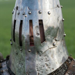 May include: A close-up of a medieval knight's helmet made of shiny, riveted metal. The helmet has vertical eye slits and is part of a suit of armor, with chainmail visible at the neck. The background is a blurred green field.