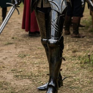 May include: Close-up of medieval armor, featuring polished black metal plates. The armor includes leg and torso protection, with intricate detailing and leather straps. A sword is held in the left hand. The scene is set outdoors on a grassy surface.