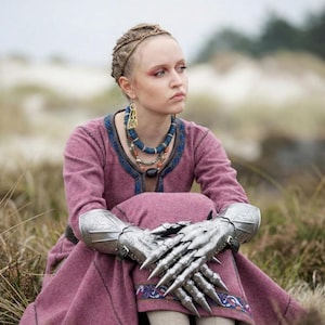 Puede incluir: Una persona con un vestido rosa con ribetes azules, peinado con trenzas y adornado con joyas elaboradas. La persona tambi&eacute;n lleva guanteletes plateados con extensiones afiladas en forma de garra. La escena est&aacute; ambientada al aire libre, entre hierba alta.