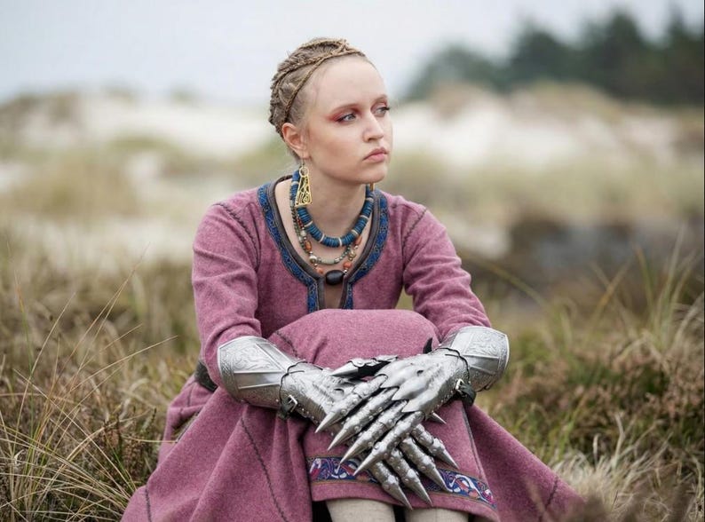 Puede incluir: Una persona con un vestido rosa con ribetes azules, peinado con trenzas y adornado con joyas elaboradas. La persona tambi&eacute;n lleva guanteletes plateados con extensiones afiladas en forma de garra. La escena est&aacute; ambientada al aire libre, entre hierba alta.