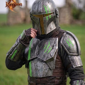 Puede incluir: Un casco y una armadura de metal desgastado con detalles verdes. El casco tiene una visera en forma de T y la armadura presenta marcas de ara&ntilde;azos. El logotipo "Keepiron" est&aacute; en la esquina superior izquierda.