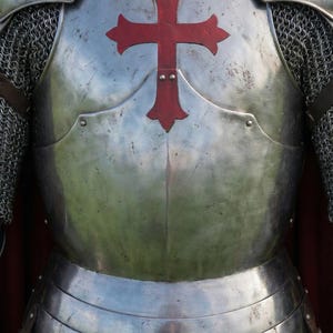 Pode incluir: Close-up de uma armadura de cavaleiro medieval. A placa peitoral de a&ccedil;o polido apresenta um grande emblema de cruz vermelha. Cota de malha e ombreiras com cruzes vermelhas tamb&eacute;m s&atilde;o vis&iacute;veis. Uma capa vermelha &eacute; parcialmente vis&iacute;vel.